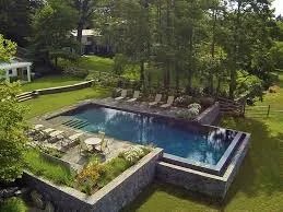 In-ground pool with dark finish, paver deck, lounge chairs, and integrated landscape lighting surrounded by mature trees