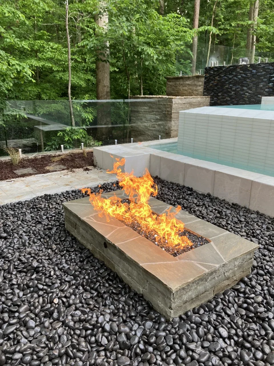 Linear concrete fire pit with flames, black river rock, pool edge, and stacked stone walls