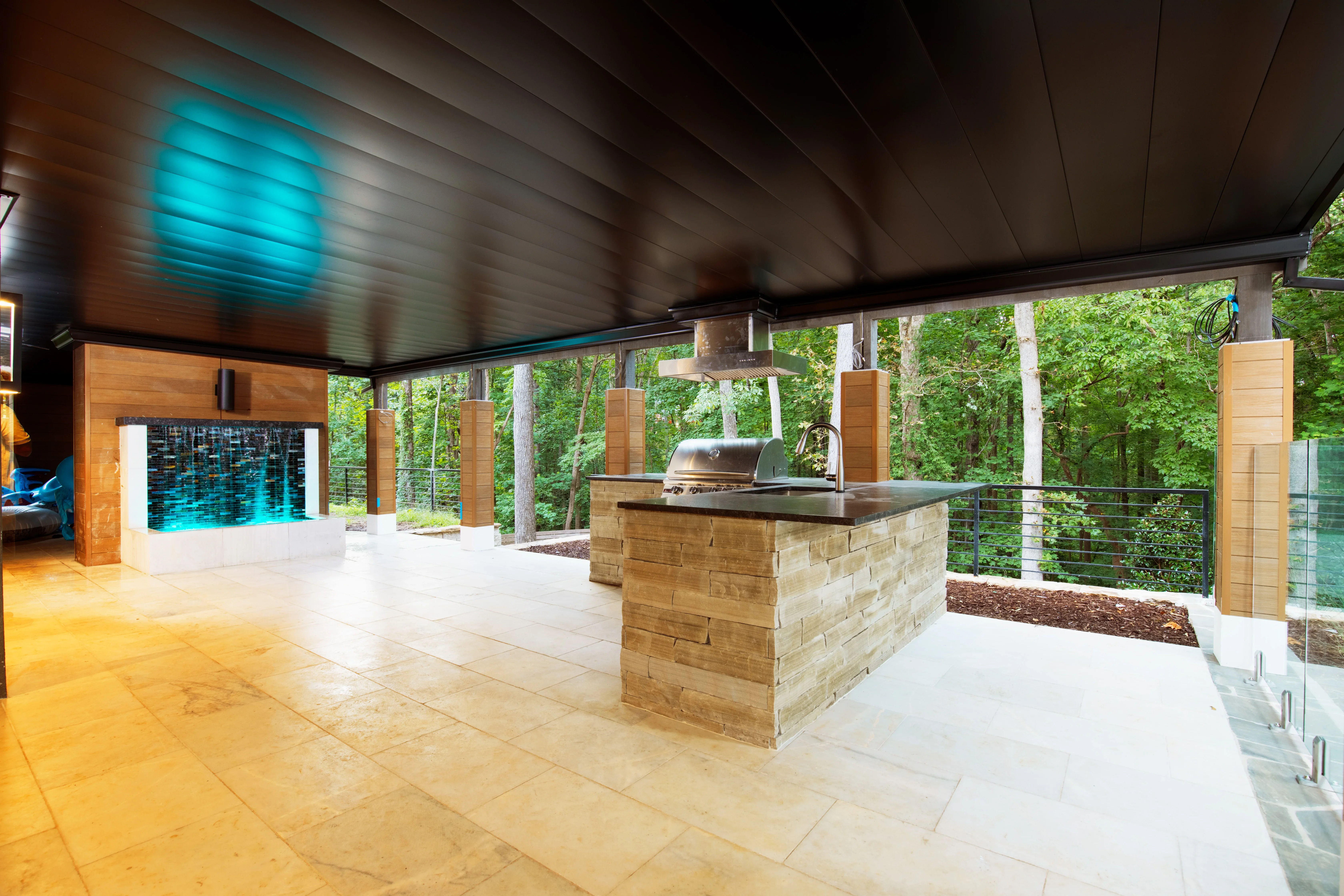 Stone island with grill, wood fireplace accent, porcelain paver patio under modern pavilion