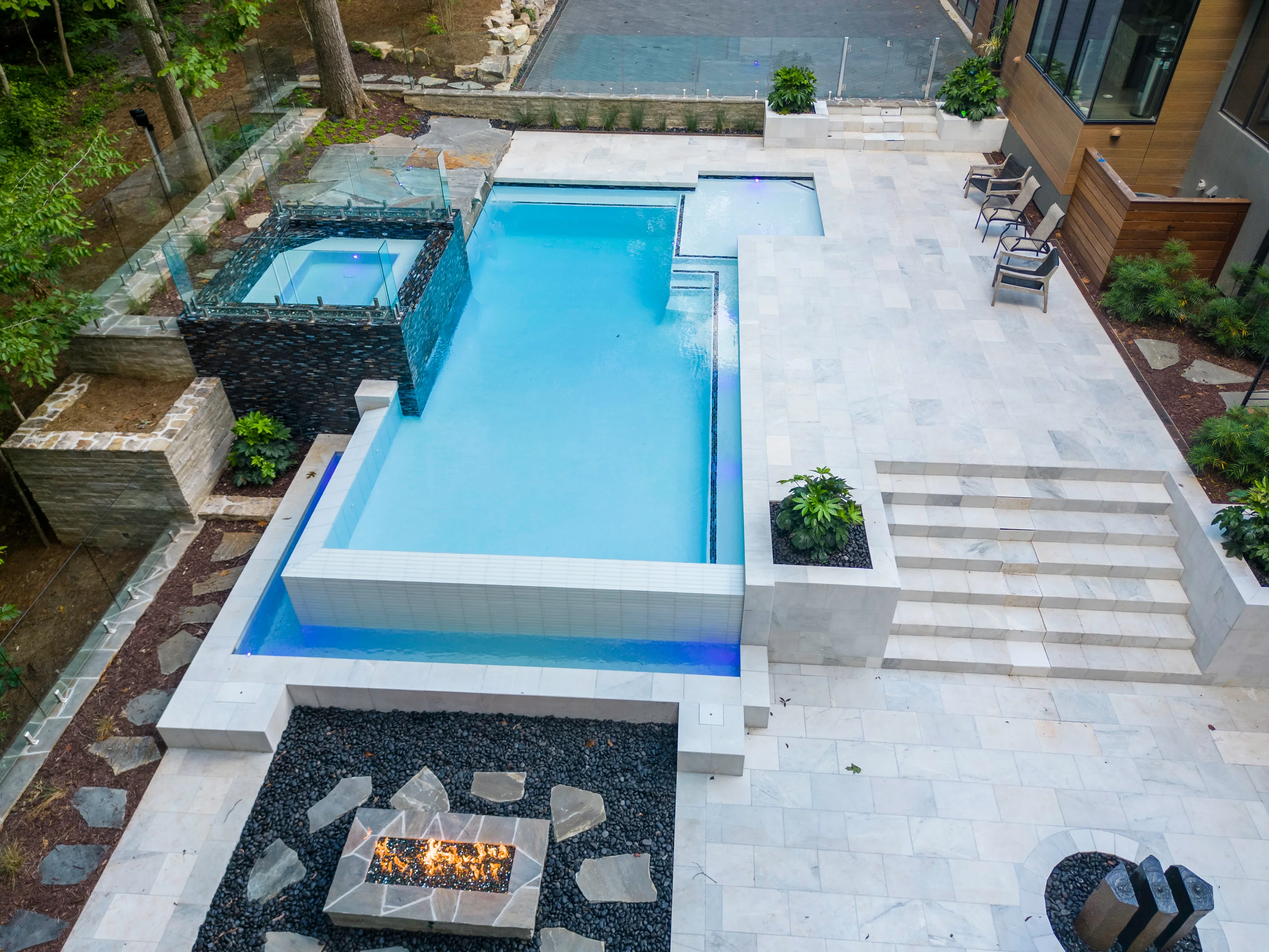 In-ground pool with white coping, blue interior, light paver patio, and dark river rock fire pit area