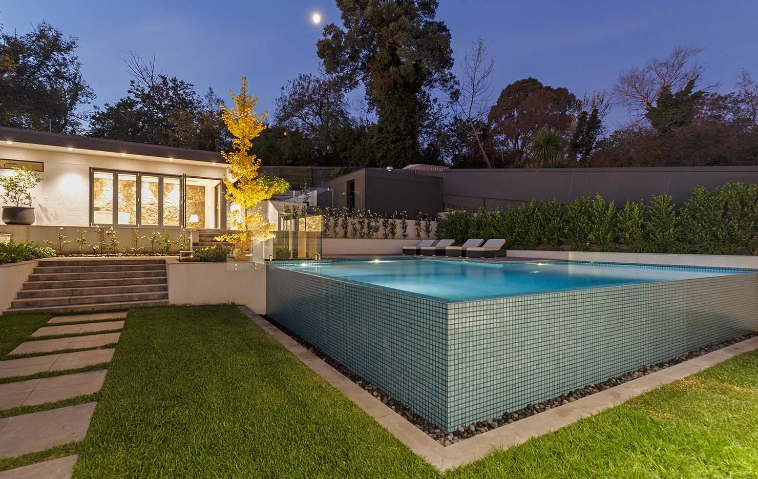 Lit swimming pool with teal tile surround, concrete deck, and modern landscape lighting at dusk