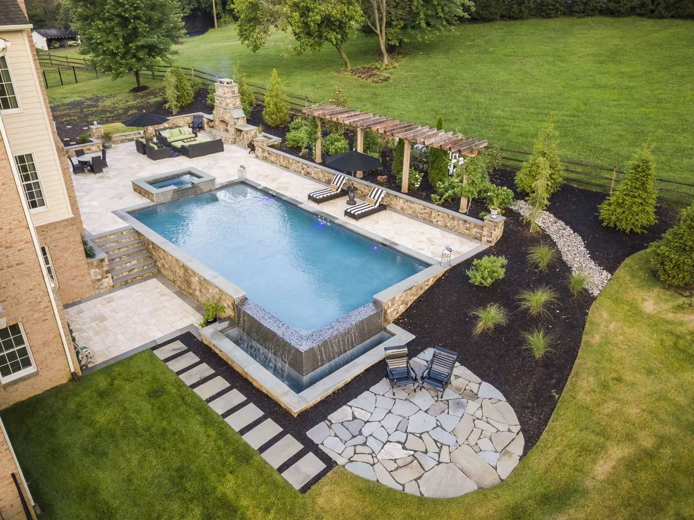 Rectangular pool with spa, travertine deck, stone walls, and wooden pergola in backyard setting
