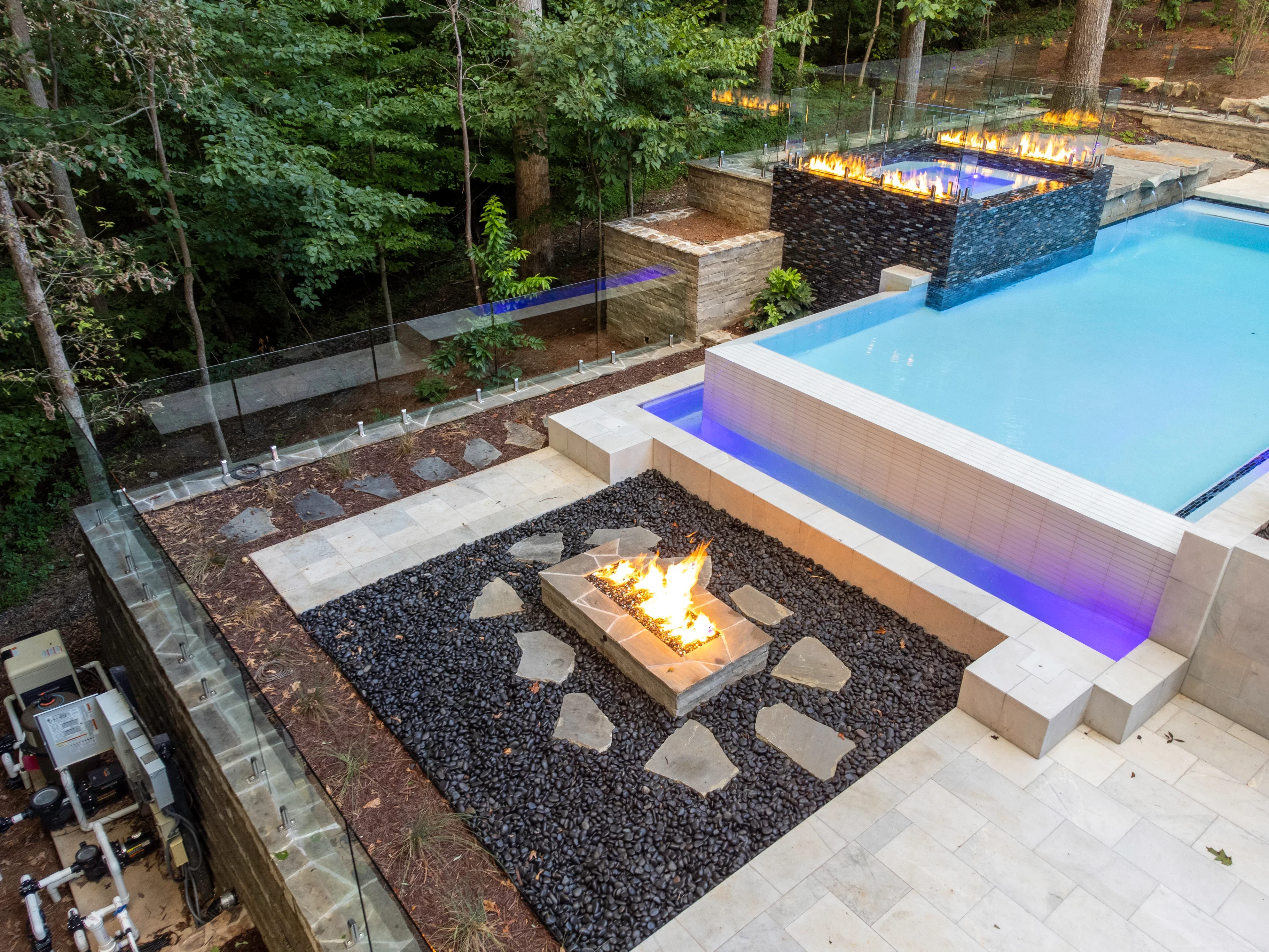 Luxury pool complex with illuminated spa, fire pit, and multi-level hardscape in wooded setting