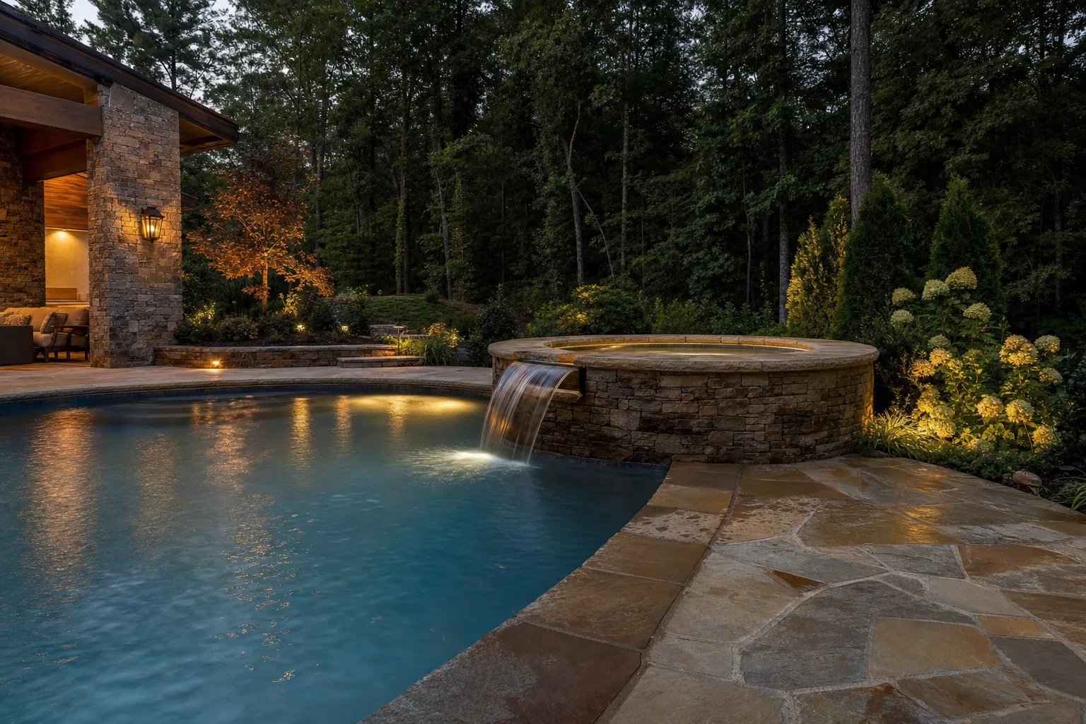 Pool with a raised stone spa spilling water into it, beside a stone patio at dusk