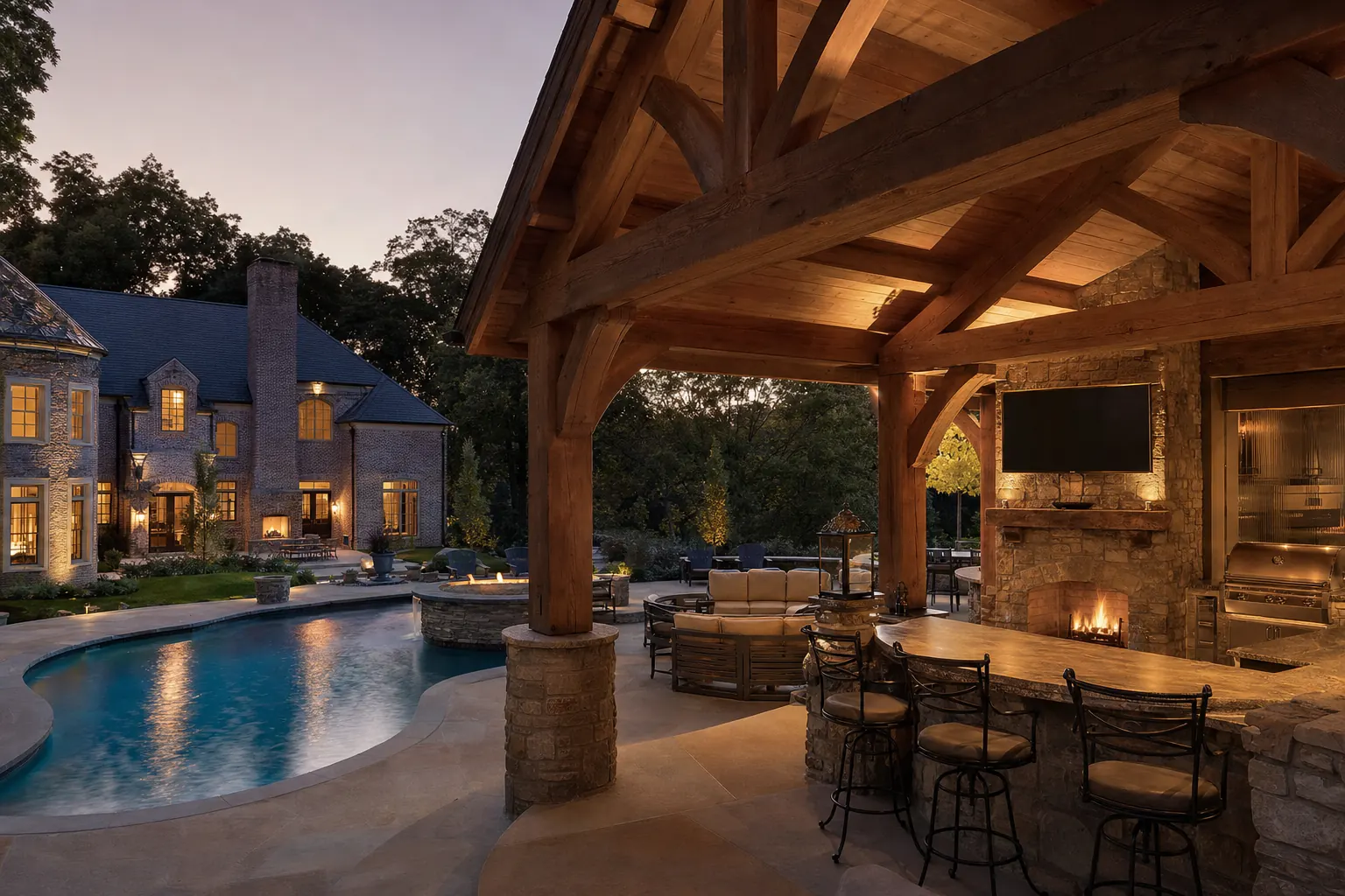 Timber pavilion with a stone outdoor kitchen, fireplace and TV beside a freeform pool at dusk