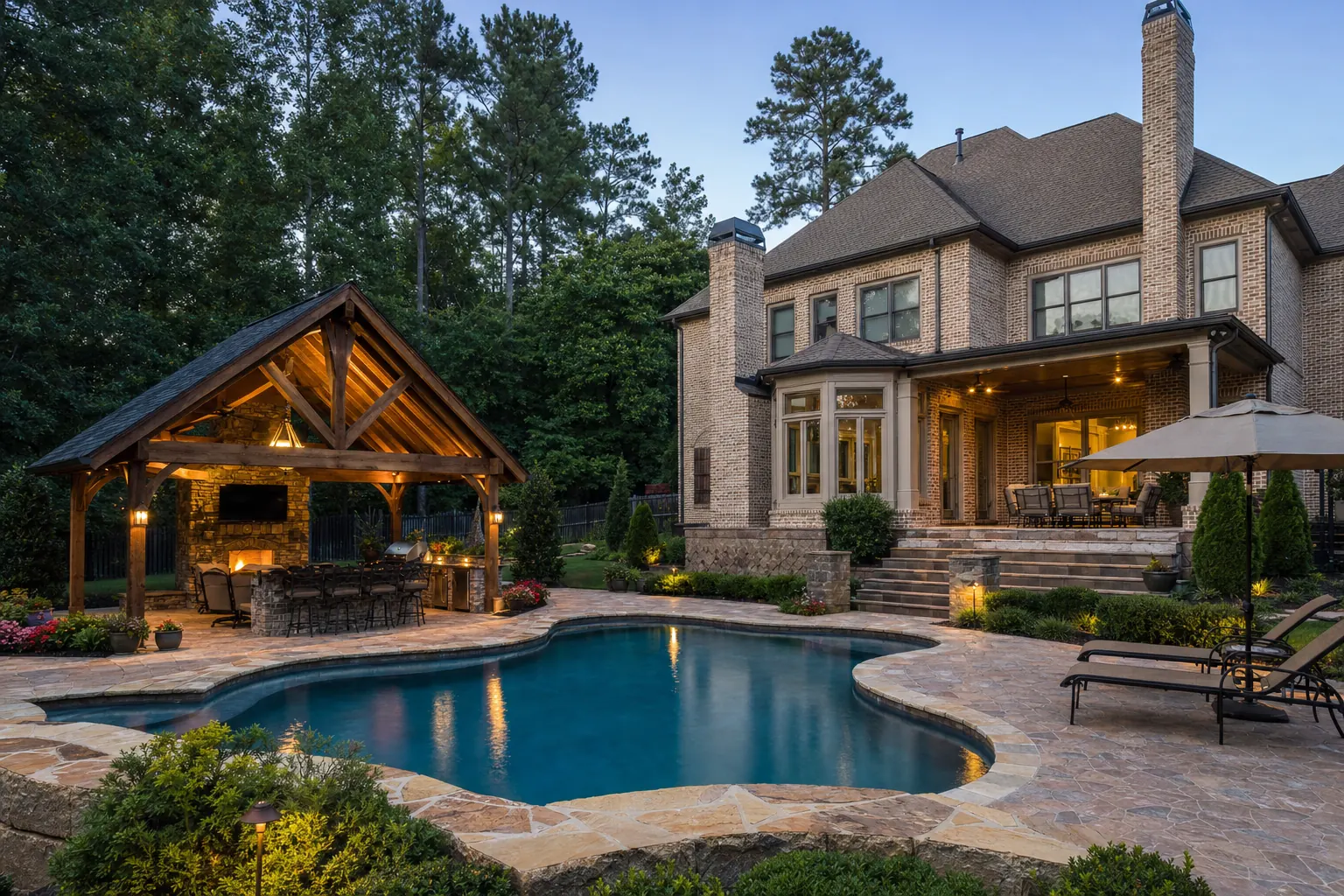 Freeform pool with a stone deck next to a timber pavilion with an outdoor kitchen and fireplace, behind a large brick home