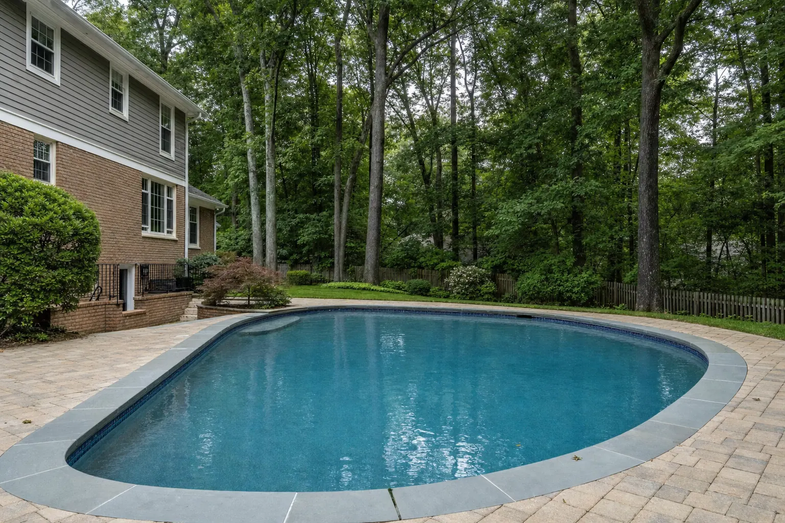 Older oval pool with new bluestone coping and a paver deck behind a two-story brick house under tall hardwoods