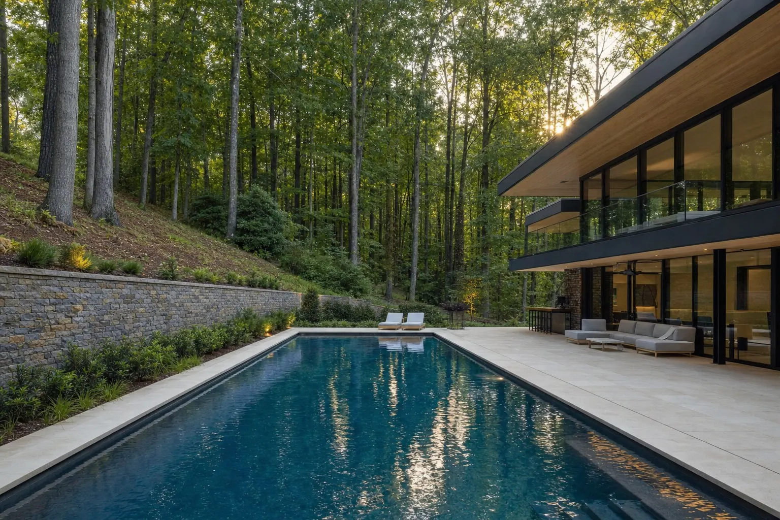 Long modern pool with a light stone deck beside a glass-walled house on a wooded slope, with a stone retaining wall uphill